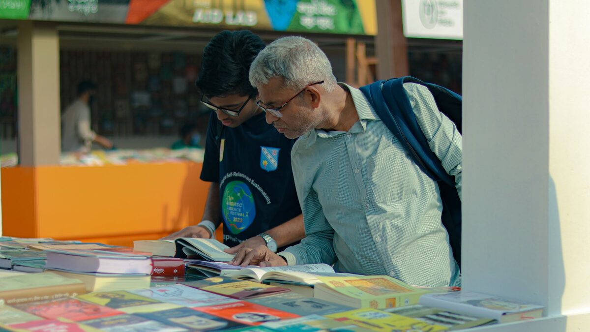 Zwei M&auml;nner bl&auml;ttern an einem Verkaufstisch im Freien in ausgelegten B&uuml;chern
