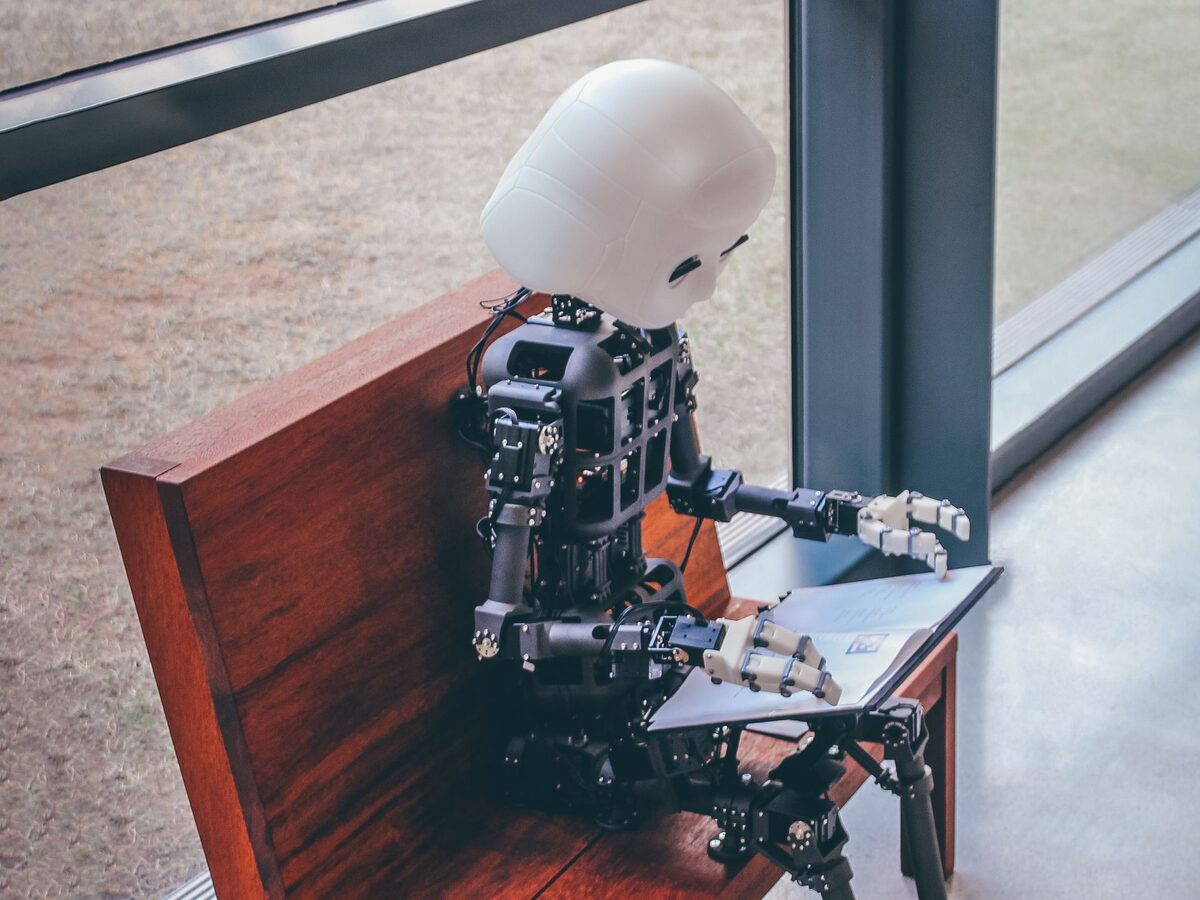 Ein kleiner humanoider Roboter sitzt auf einer Holzbank und bl&auml;ttert in einem Buch
