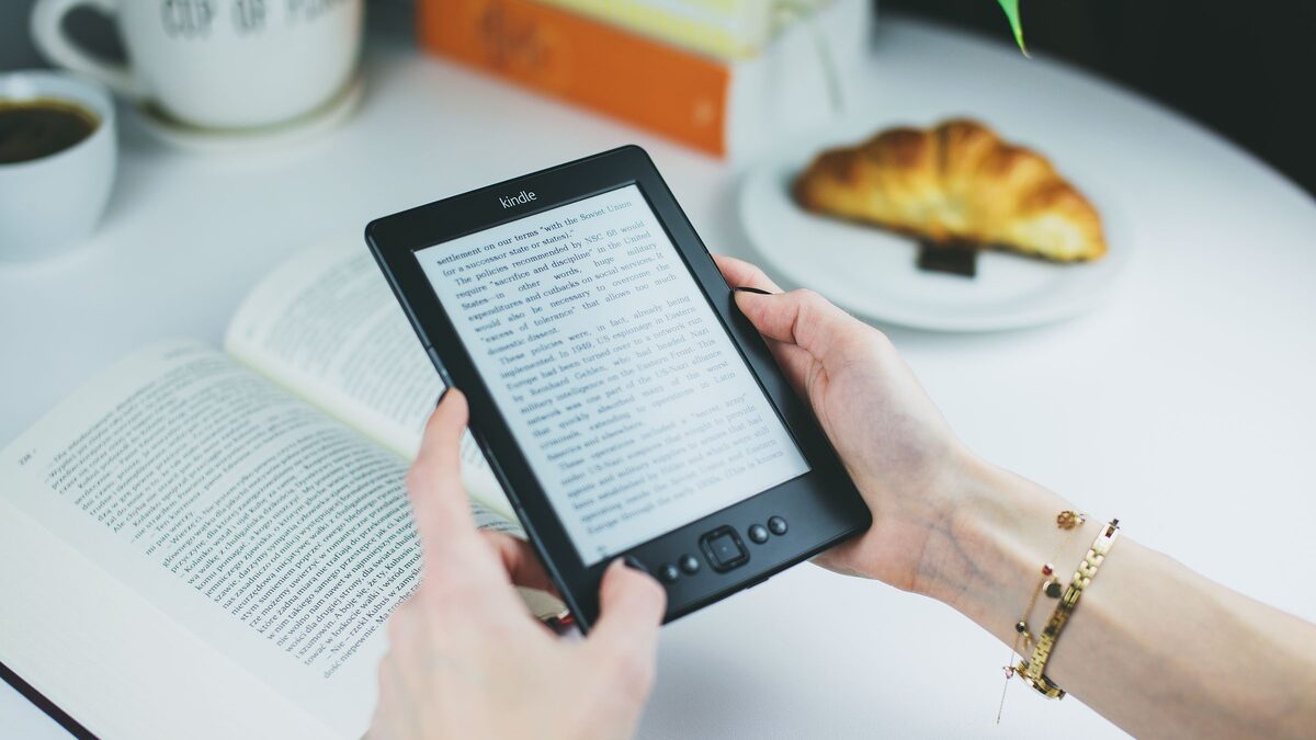 Eine Frau h&auml;lt einen Kindle in den H&auml;nden, auf dem Tisch davor liegen ein Buch und ein Croissant