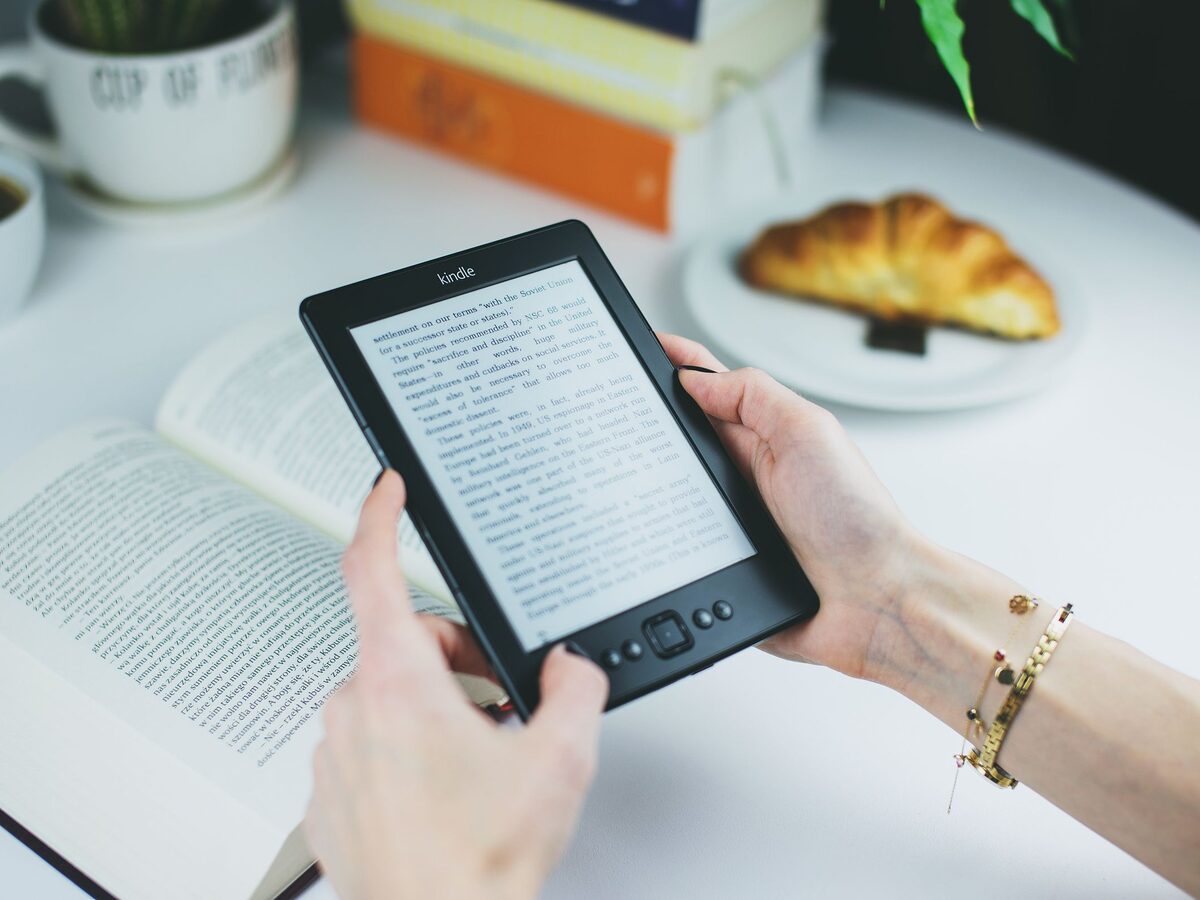 Eine Frau h&auml;lt einen Kindle in den H&auml;nden, auf dem Tisch davor liegen ein Buch und ein Croissant
