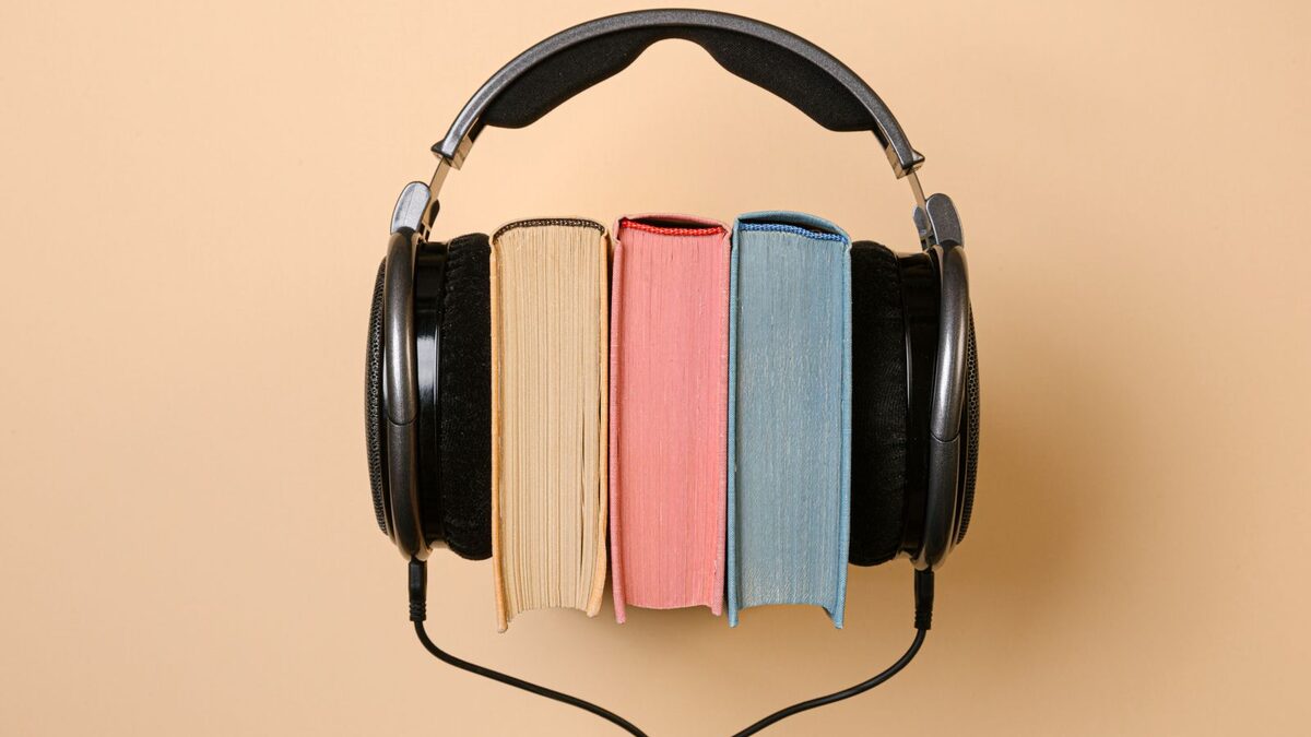 Ein schwarzer Kopfh&ouml;rer umschlie&szlig;t drei B&uuml;cher mit farbigem Schnitt vor beigem Hintergrund