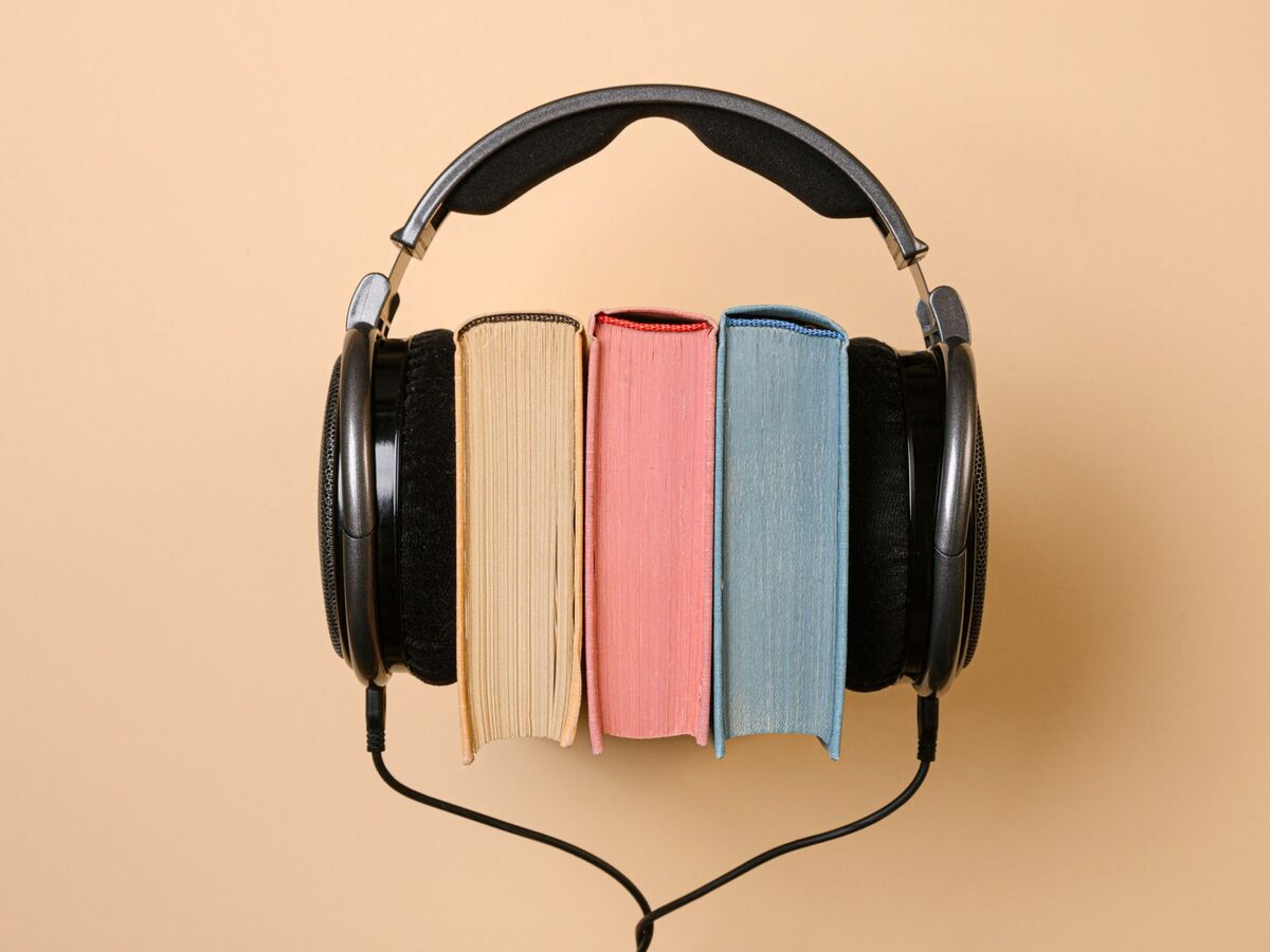 Ein schwarzer Kopfh&ouml;rer umschlie&szlig;t drei B&uuml;cher mit farbigem Schnitt vor beigem Hintergrund
